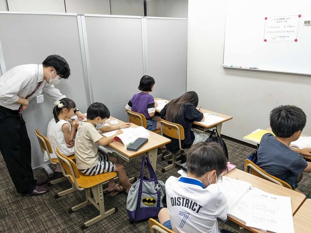 教室で数理パズルの教材に取り組む子どもたちと、机を回って指導する講師